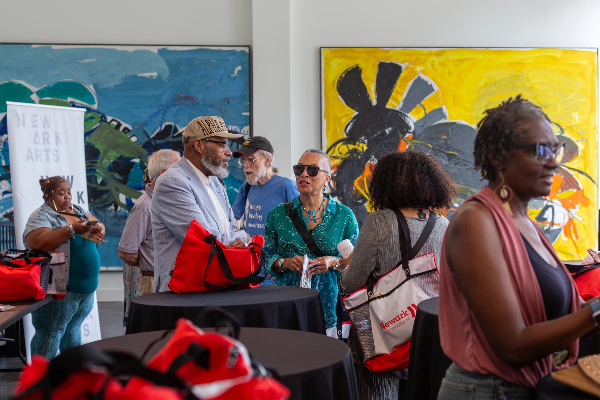 Attendees mingle at a Newark Arts gallery reception, holding red and Newark Happening tote bags, with a "Newark Arts" banner on the left and two large abstract paintings in blue and yellow hanging on the wall behind them.