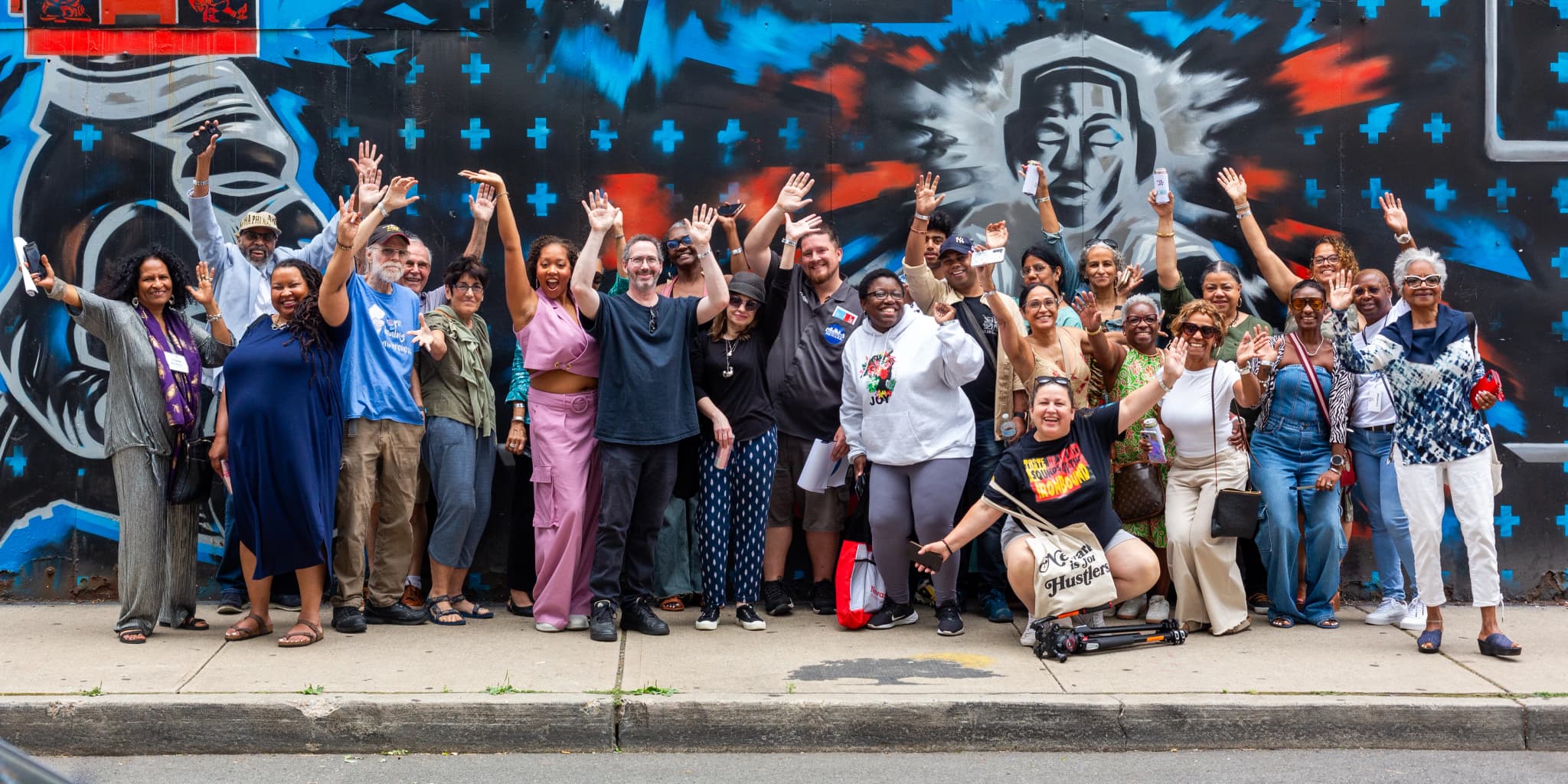 A large group of roughly thirty people stand on a sidewalk with arms raised and cheering, posed in front of a striking blue, black, and red street mural depicting two robed figures with patterned cross motifs.