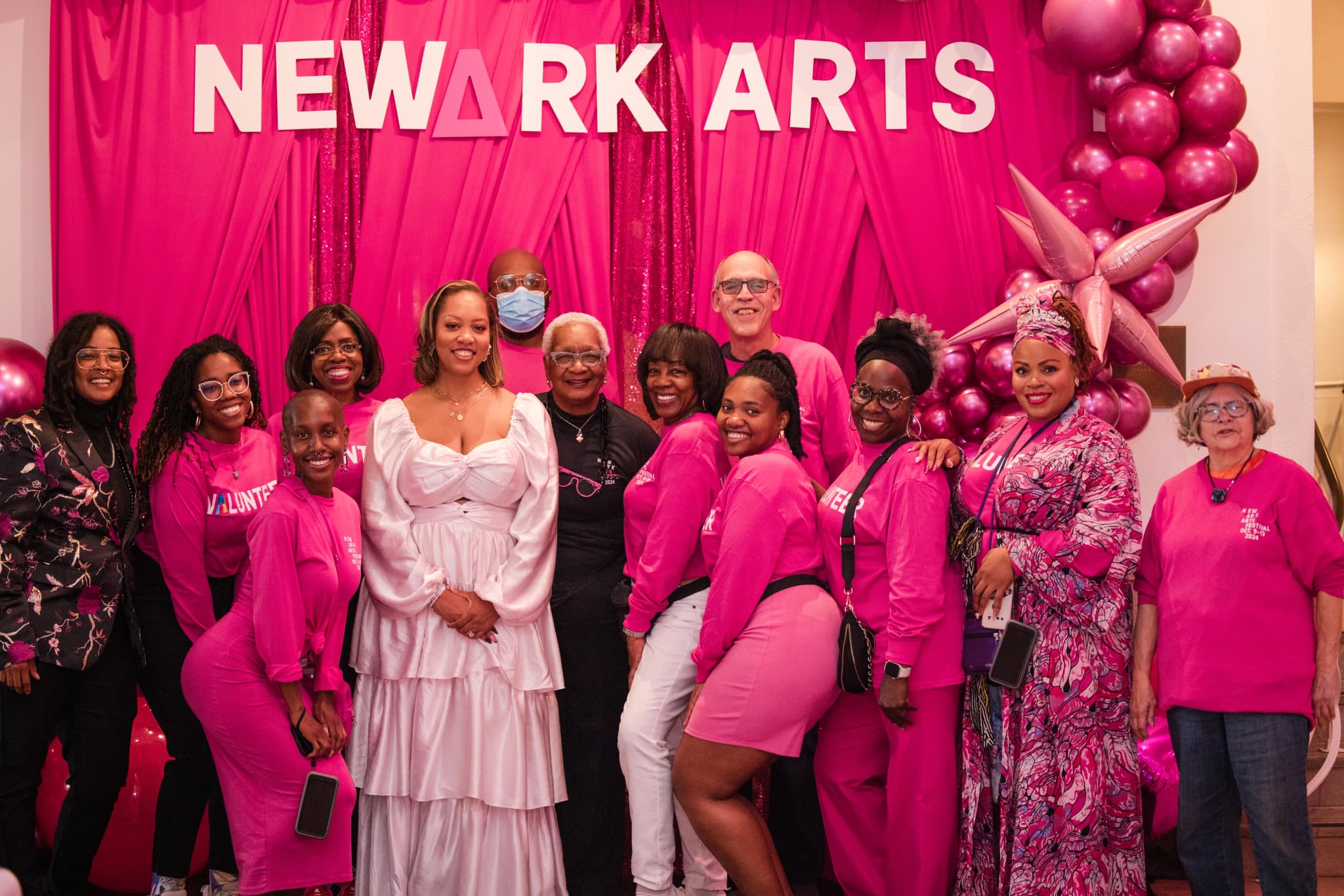 A group of about a dozen Newark Arts volunteers and supporters, most wearing bright pink shirts and outfits, smile and pose together in front of a hot-pink curtain backdrop with large white "NEWARK ARTS" lettering and a cluster of metallic pink balloons