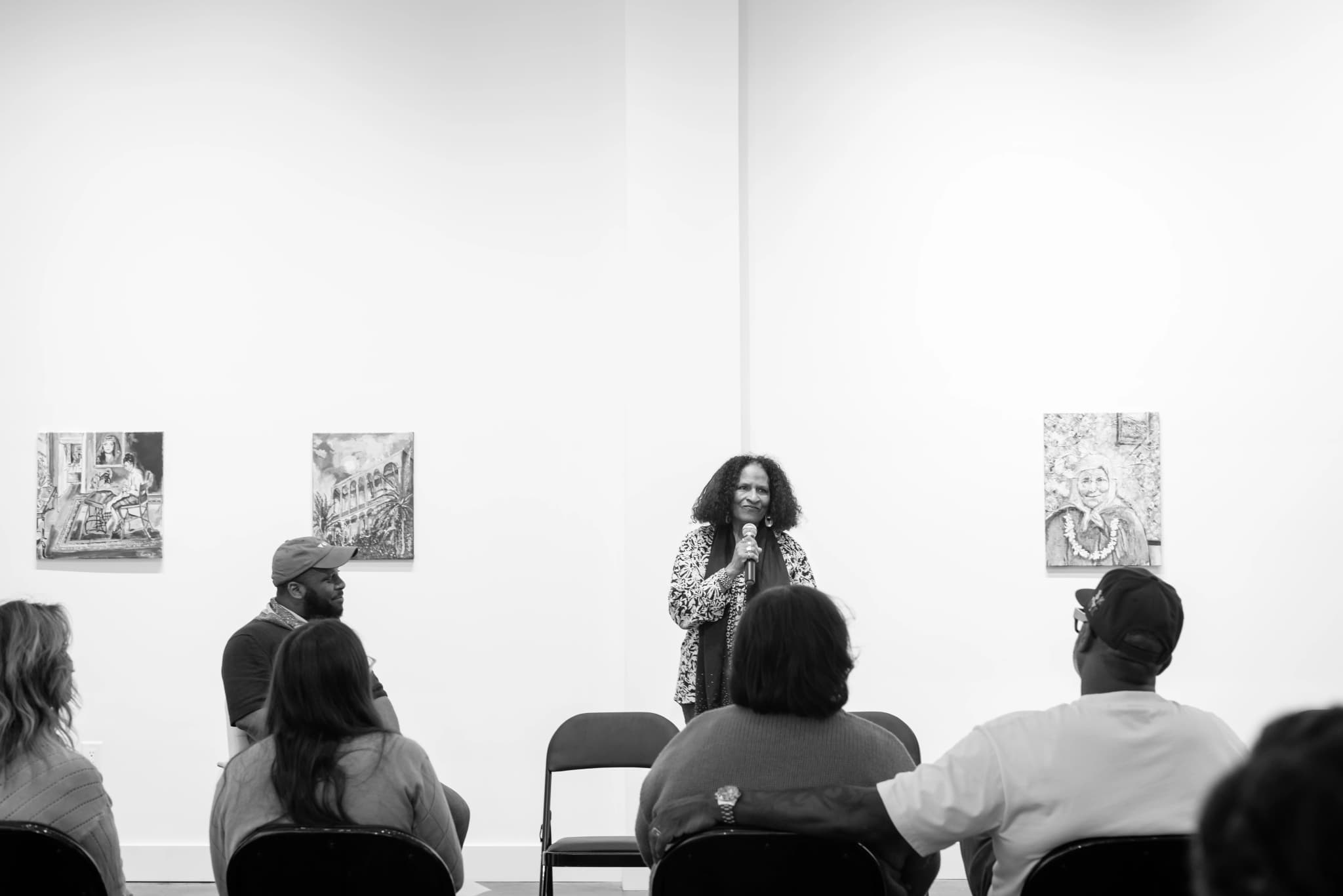 Black-and-white photo of a woman with curly hair, wearing a patterned jacket and scarf, speaking into a microphone in a bright gallery space, with three small paintings on the wall behind her and audience members seated in the foreground