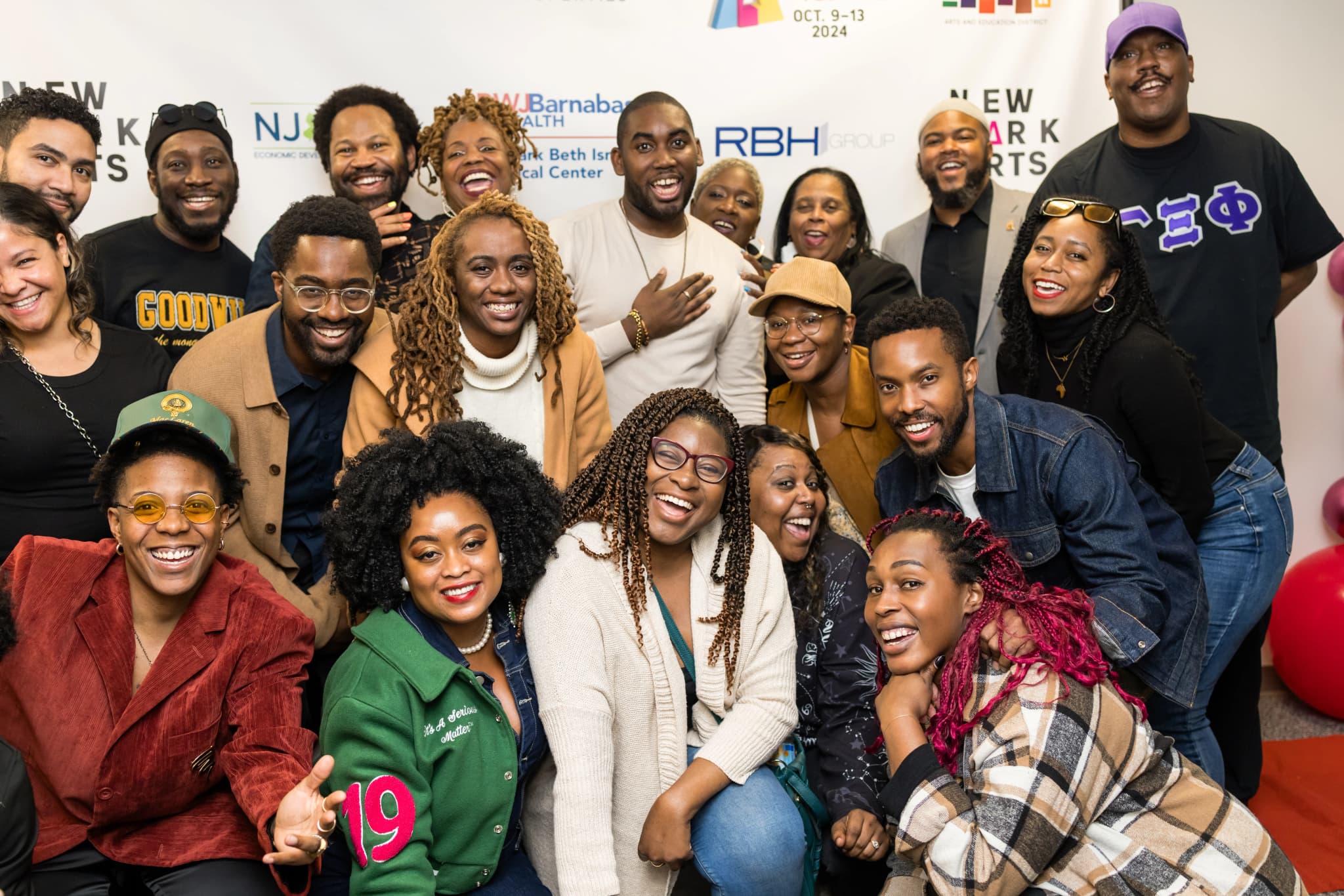 A large group of roughly twenty attendees smiling and laughing together for a group photo at the Newark Arts Festival, posed in front of a sponsor backdrop featuring logos for RWJBarnabas Health, NJ Economic Development, Newark Beth Israel Medical Center, and RBH Group