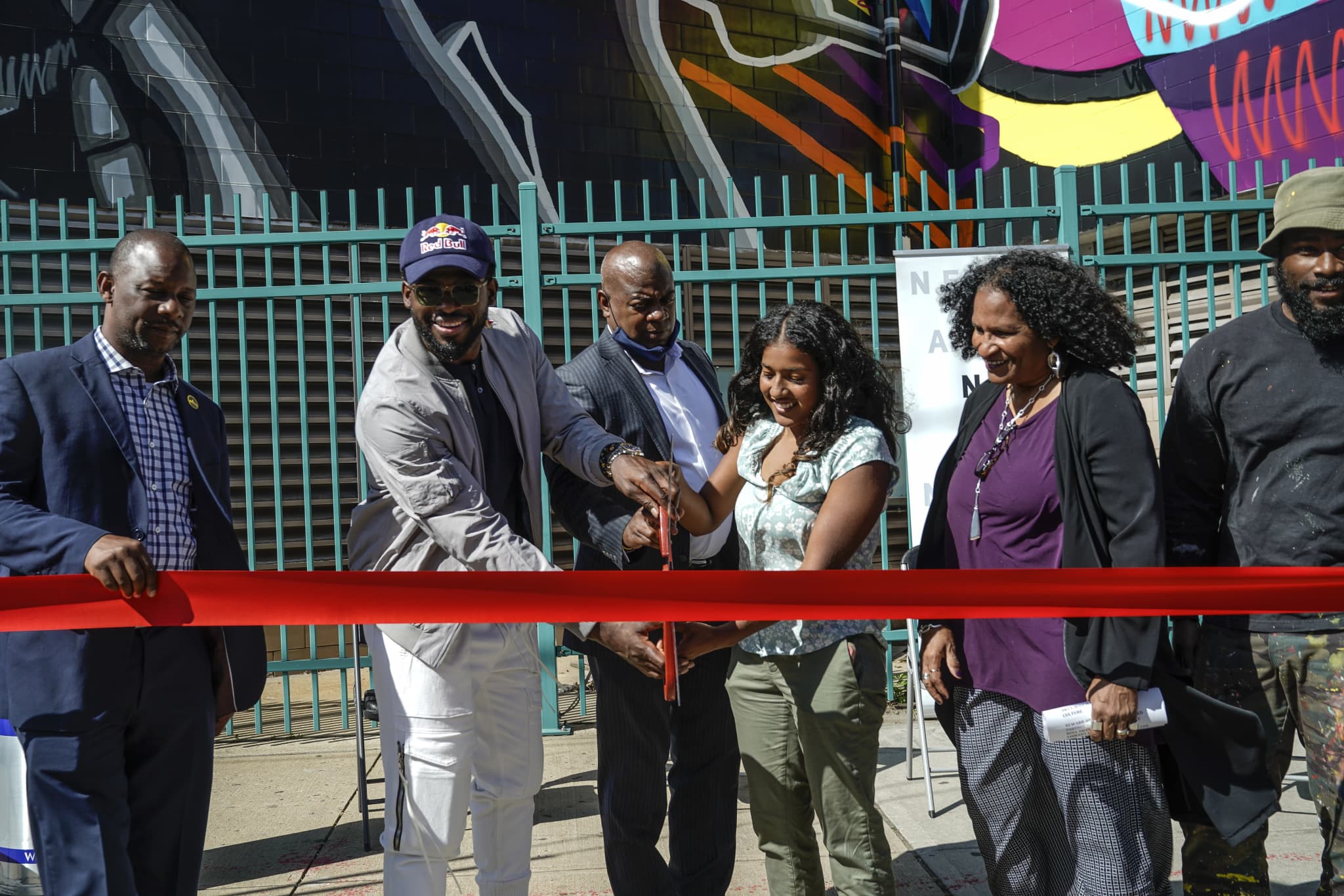 A ribbon-cutting ceremony in front of a teal metal fence at the P.K. Subban mural site, with six people gathered around a long red ribbon. A man in a Red Bull cap and gray bomber jacket, a man in a pinstriped blazer, and a smiling young woman in a floral blouse hold large scissors together, while others stand beside them looking on. The colorful graffiti mural is visible on the wall behind them