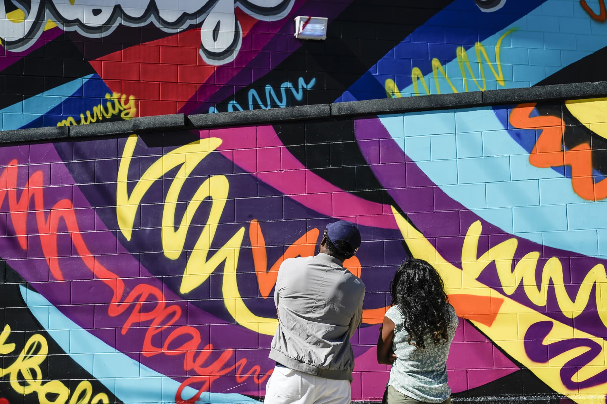 Rear view of a man and young woman standing close together, looking up at the colorful cinder-block mural covered in swirling brushstrokes and graffiti-style lettering, with the words "community" and "play" partially visible among the designs.